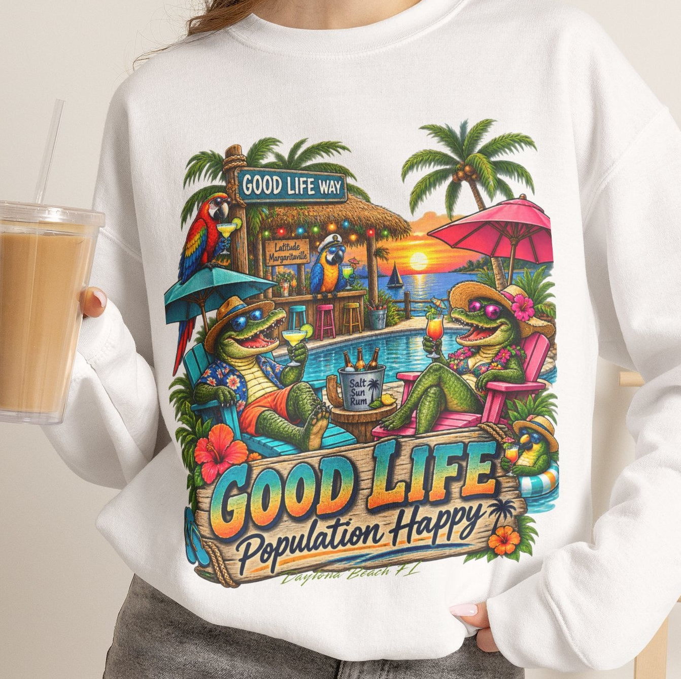 A woman wearing a white crewneck sweatshirt with a colorful graphic print of a tropical frog bar scene and the text 'GOOD LIFE Population Happy'. She is holding an iced coffee and wearing gray denim shorts in a brightly lit indoor setting.