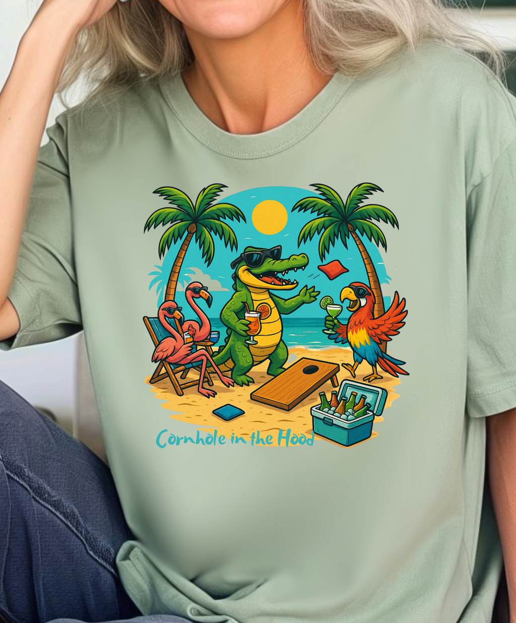 Person wearing a light green t-shirt with a colorful graphic of a crocodile, flamingos, and a parrot playing cornhole.