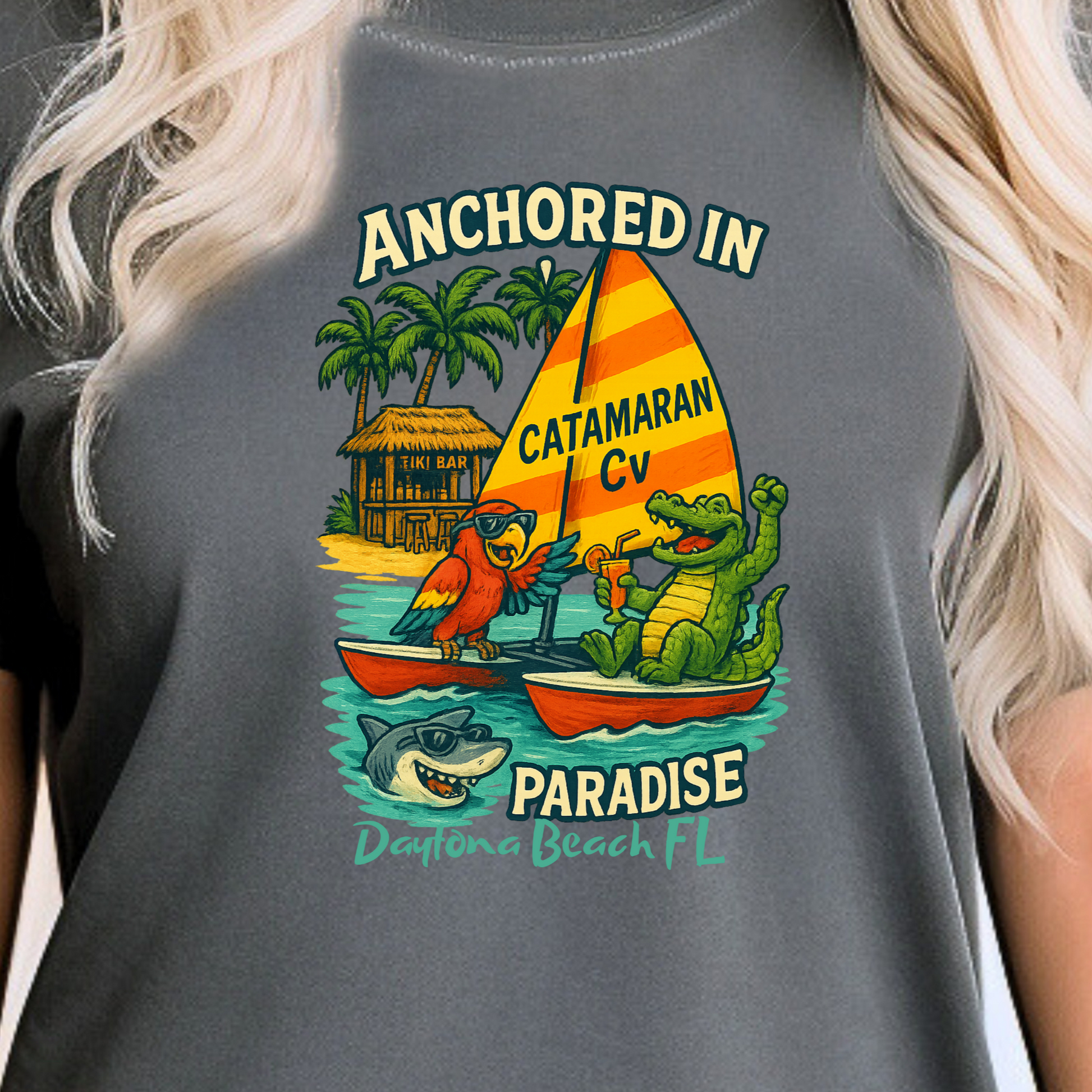 Person wearing a gray t-shirt with a colorful graphic design featuring a catamaran, crocodile, and palm trees.