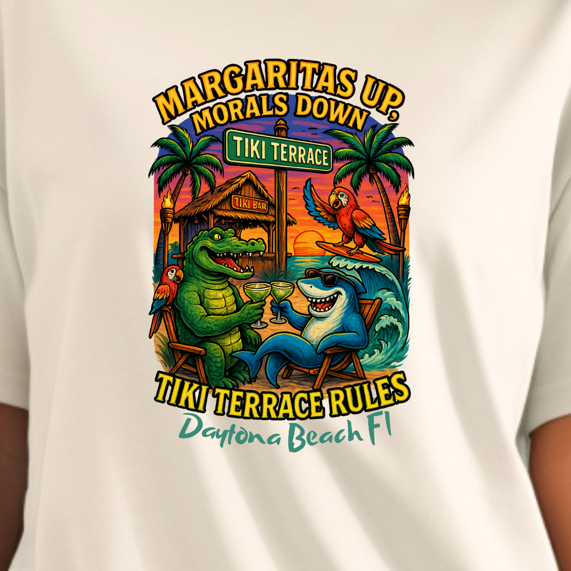 White t-shirt with a colorful graphic design featuring a crocodile and shark, text 'Margaritas Up Morals Down Tiki Terrace' on a beach background.