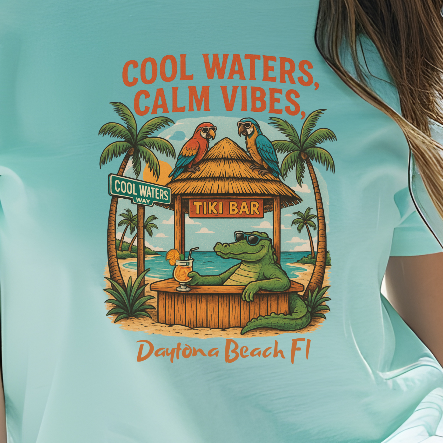 Person wearing a light blue t-shirt with a tropical design and text, standing on a beach.